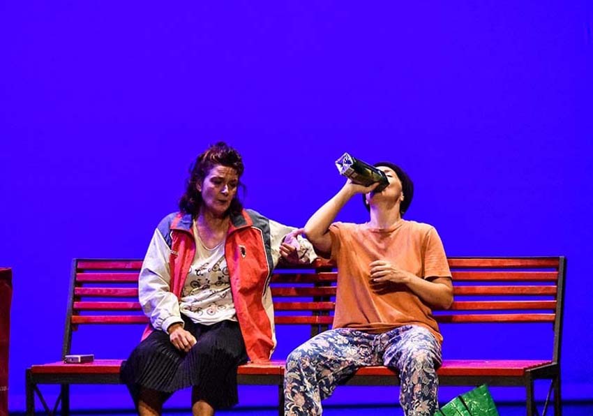 Two actresses sitting on a bench one of them drinking